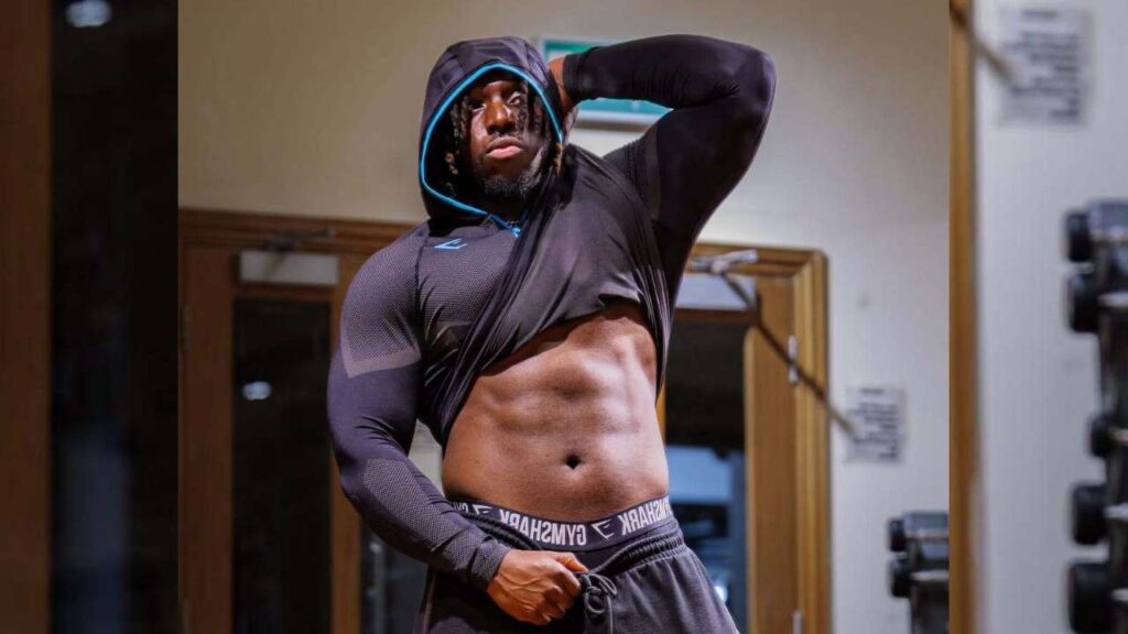 Nonso Chinye in a dark athletic hoodie with blue trim lifts his shirt to reveal abs, posing in a gym with a rack.