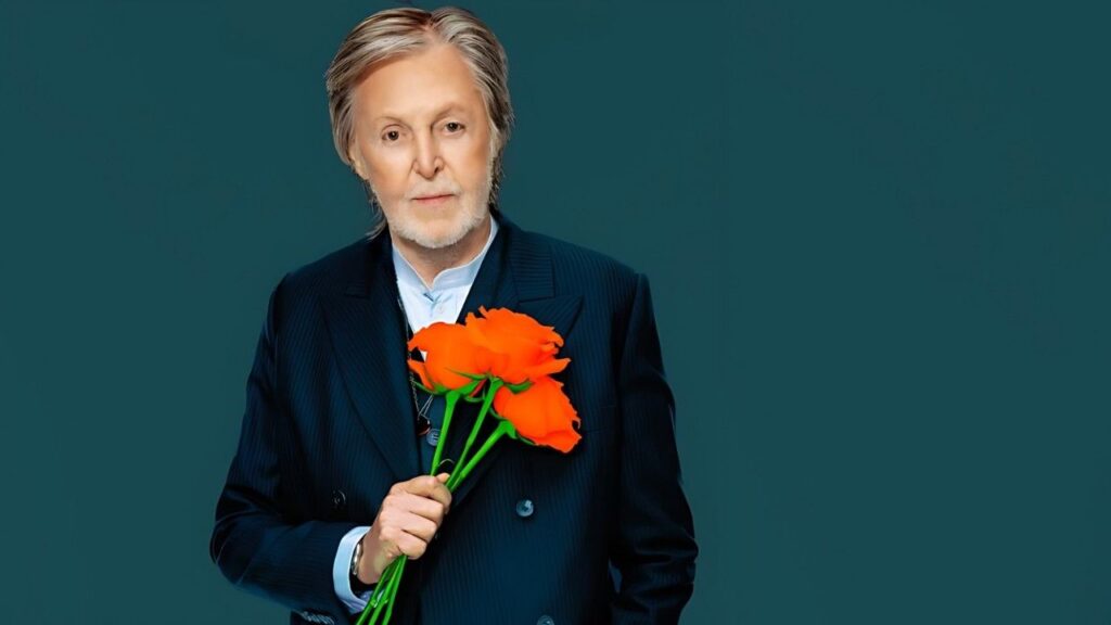 Paul McCartney with gray hair in a navy suit holds a bouquet of bright orange roses, posed against a solid teal background.