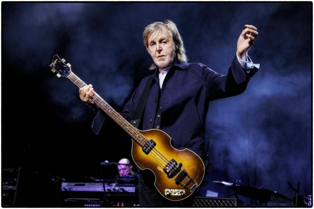 Paul McCartney onstage with a sunburst bass, left hand on the neck and right hand raised, smoky blue spotlight behind him.