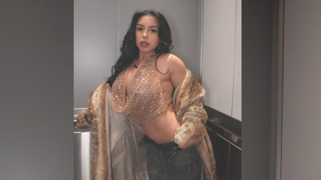 Priscila Montoya with long dark hair and bold makeup poses in an elevator, wearing a sparkling nude-beige halter top and a fur coat draped over her shoulders.