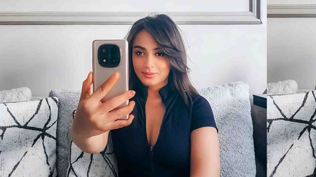 Rim Fethi snaps a mirror selfie on a gray sofa, holding a large smartphone, wearing a black top, with patterned cushions.