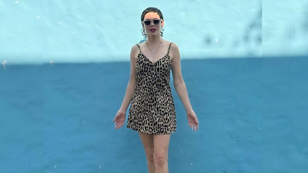 Rose McGowan Leopard-print mini dress with dark sunglasses and a necklace, standing before a two-tone blue wall with arms at her sides.