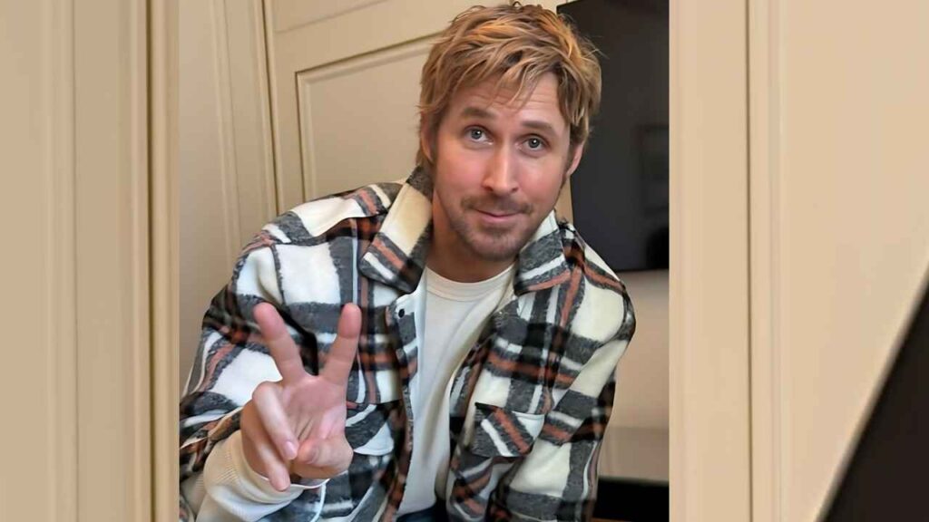 Ryan Gosling with tousled hair in a white tee and plaid flannel, leaning in a doorway and flashing peace sign with a warm smile.