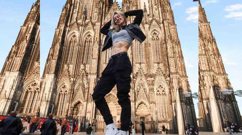 Sabrina Cesta in a crop top, black cargo pants, white sneakers poses with hands behind her head in front of a towering Gothic cathedral at sunset.