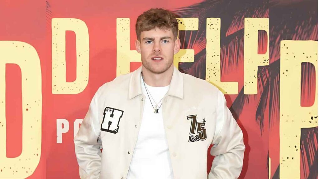Sam Goatlifters poses in front of a red poster with yellow letters, wearing a light beige jacket with patches over a white tee.