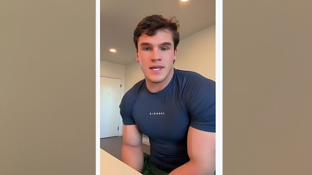 Samuel Millsap with short brown hair in a tight blue athletic shirt leans toward the camera in a bright, simple room, smiling.