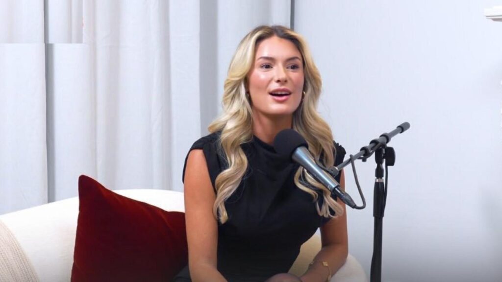 Shelby Haas in a black sleeveless top sits on a beige sofa with a red cushion, speaking into a silver microphone on a stand.