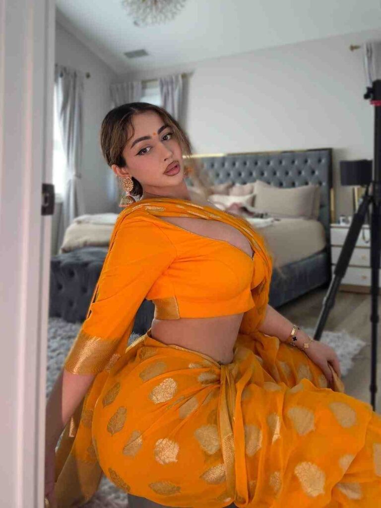 Shilpa Sethi photo in a bright orange saree with gold accents poses on a tufted bed in a modern bedroom, wearing gold earrings.