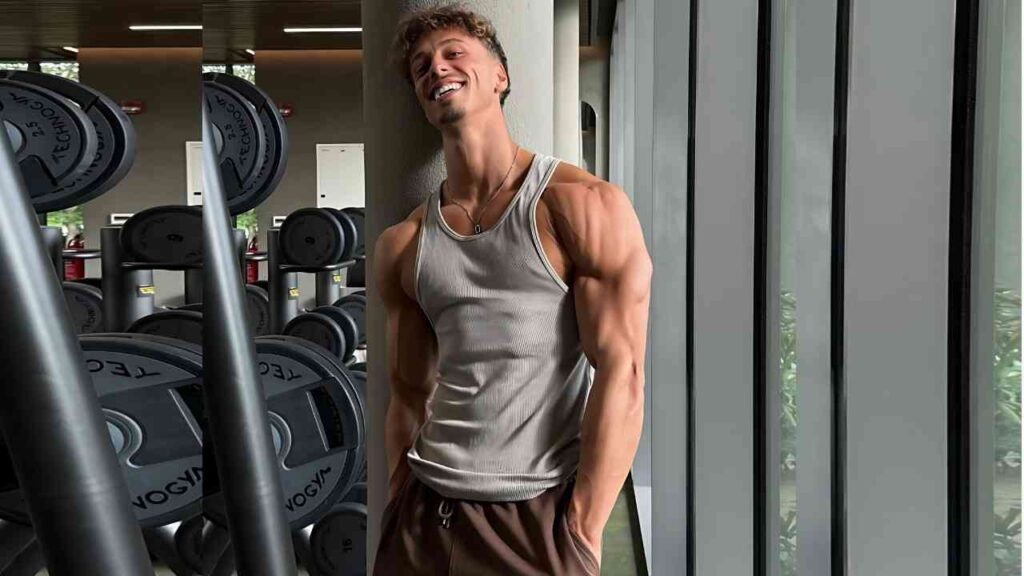 Soufiane Haloui in a gray sleeveless shirt and brown shorts leans against a pillar in a bright gym, smiling amid stacked weights.