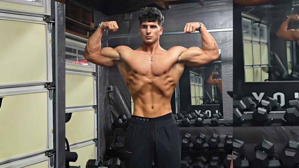 Stef Meyers shirtless in a gym flexes his biceps, displaying defined abs in black shorts, beside dumbbells and mirrors.