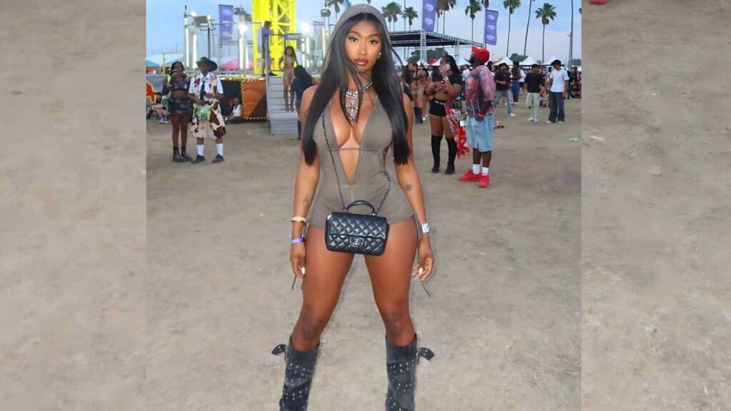 Tavi Larai in taupe plunging romper and knee-high boots poses with a small black quilted handbag at a sandy festival, palm trees.