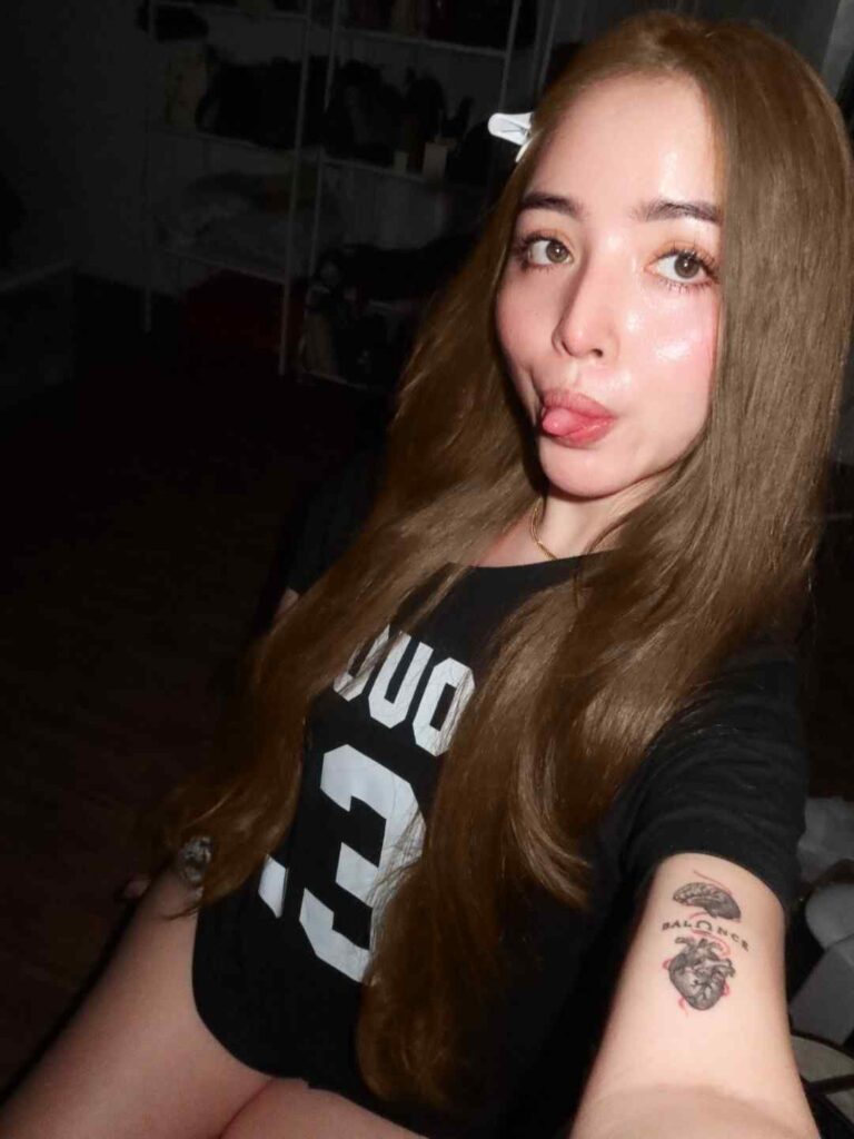 natalyn credo peralta biography with long brown hair and a white clip, wearing a black graphic tee; forearm tattoo visible in a dim room.