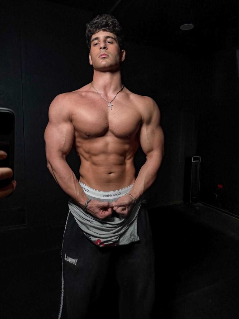 stef meyers tiktok in a dark room flexes abs and biceps; CK waistband shows over gray sweats, silver cross necklace.