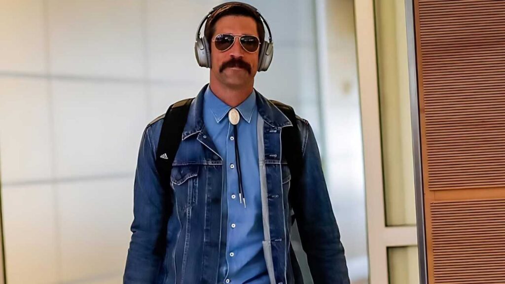 Aaron Rodgers in aviator sunglasses, blue shirt with bolo tie, denim jacket, headphones, and backpack in a bright hallway.
