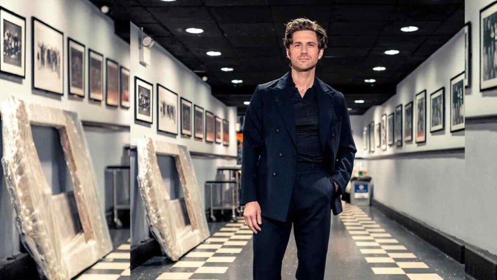 Aaron Tveit in a dark navy suit stands in a gallery hallway, framed photos along the walls and frames leaning on a checkered floor.