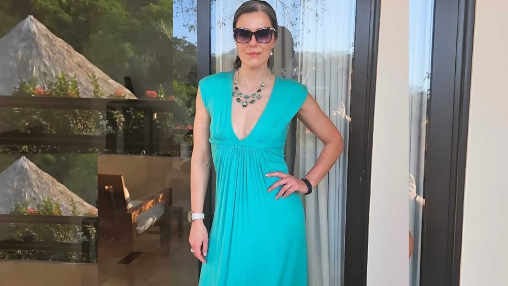 Adrianne Curry with a bold green necklace, sunglasses, and white watch, posing on a balcony.