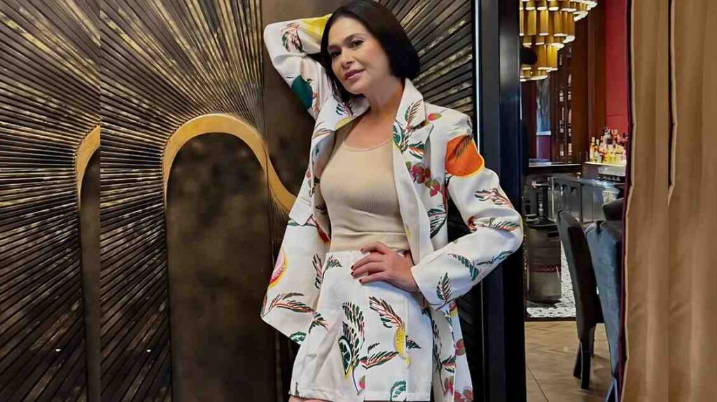 Aiko Melendez in white blazer and shorts with colorful leaf print, beige top, posing with hand on hip, against a wood-panel wall.