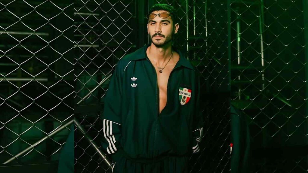 Alejandro Speitzer in a dark green Adidas tracksuit leans against a chain-link fence, glasses on his head, chest exposed with necklace.