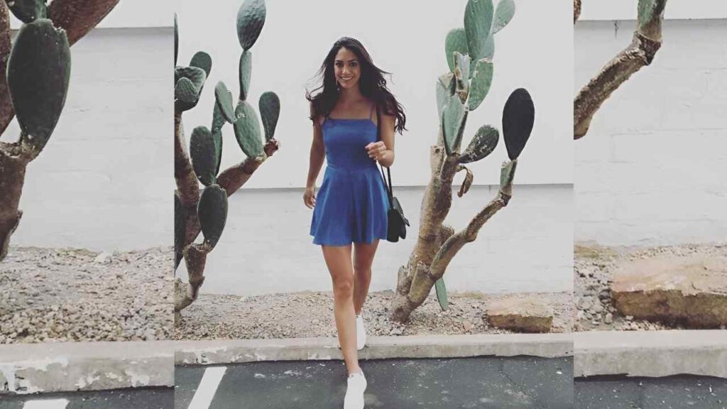 Allison Stokke in a blue dress walks between large cactus plants against a white wall, carrying a small black bag and smiling.