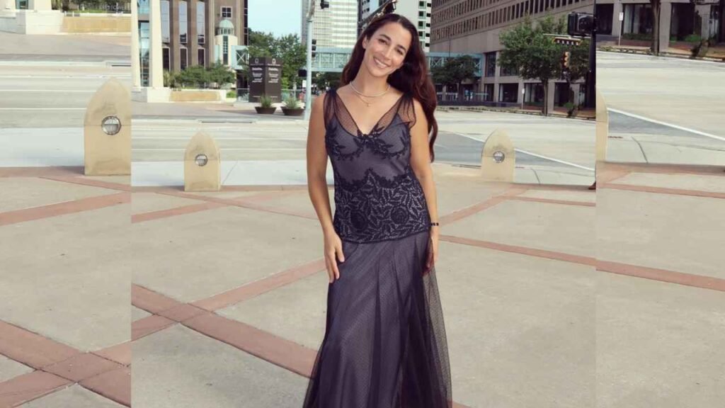 Aly Raisman in a black lace evening gown posing in a sunny urban plaza with modern office buildings in the background.