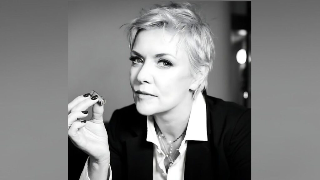 Amanda Tapping with short platinum hair in a dark blazer, holding a small object between her fingers and gazing at the camera.