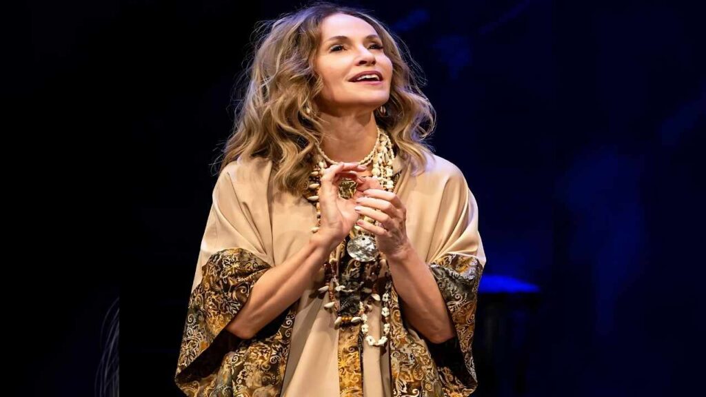 Amy Brenneman with shoulder-length wavy blonde hair on a dark stage, wearing a beige robe with ornate cuffs, clasping her hands.