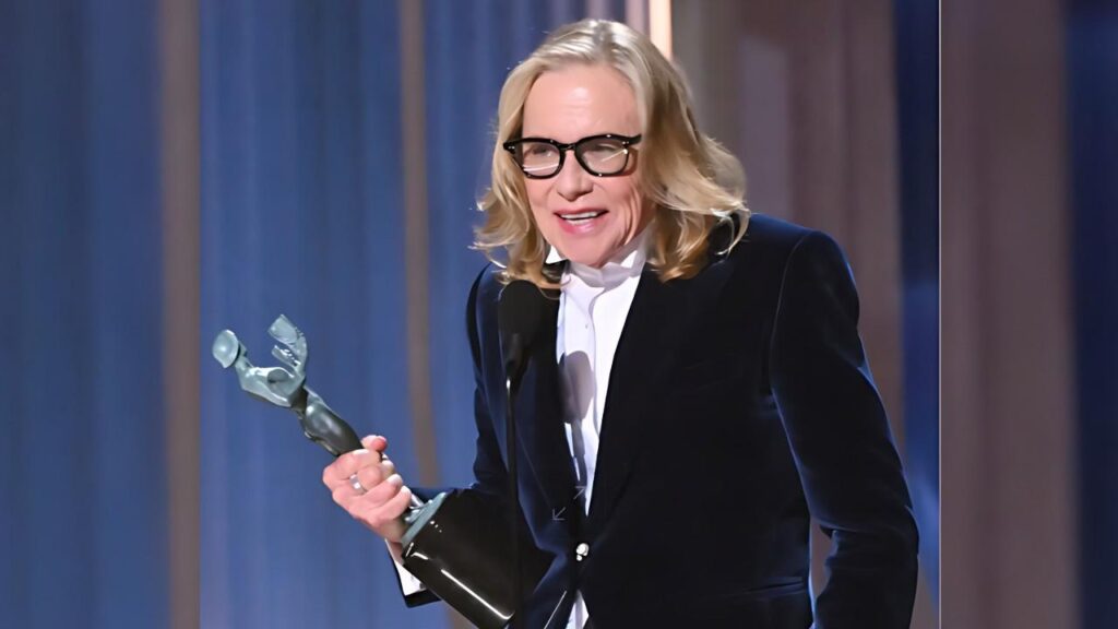Amy Madigan in velvet blazer and white shirt, wearing glasses, smiles while holding a silver trophy at a microphone on stage with blue drapes.