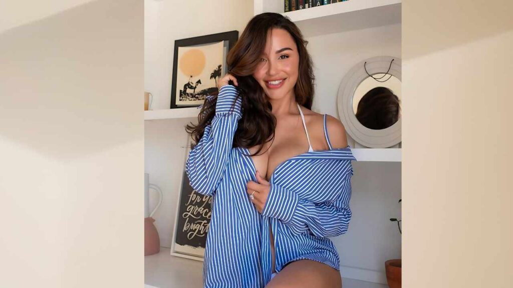 Ana Cheri in a blue-and-white striped off-shoulder shirt leans against a shelf, with round mirror and framed art behind.