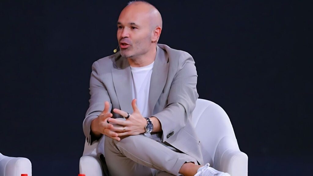 Andrés Iniesta in a light gray suit and white tee sits on a white chair, gesturing with his hands while speaking on stage.