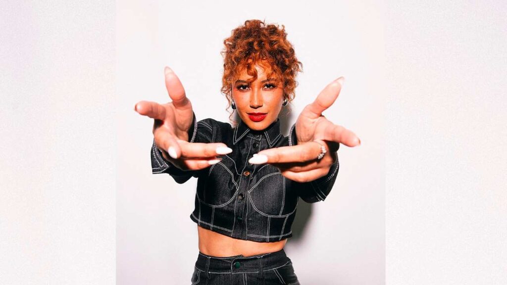 Andy Allo with curly red hair in a cropped dark denim top, reaching toward the camera with both hands.