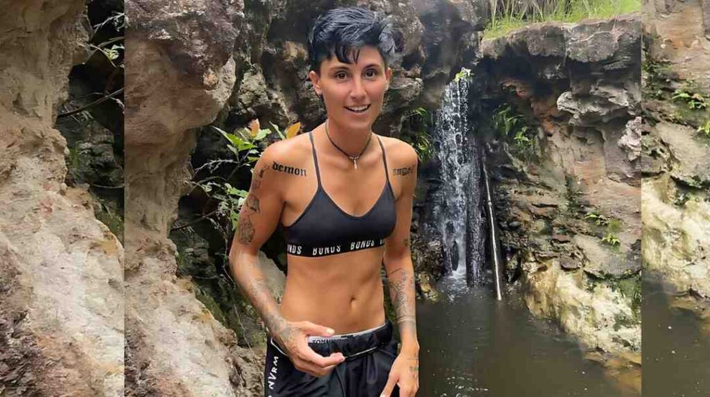 Angela Taylor with short dark hair in a black sports bra and shorts stands by a rocky waterfall and pool, tattoos on arms.