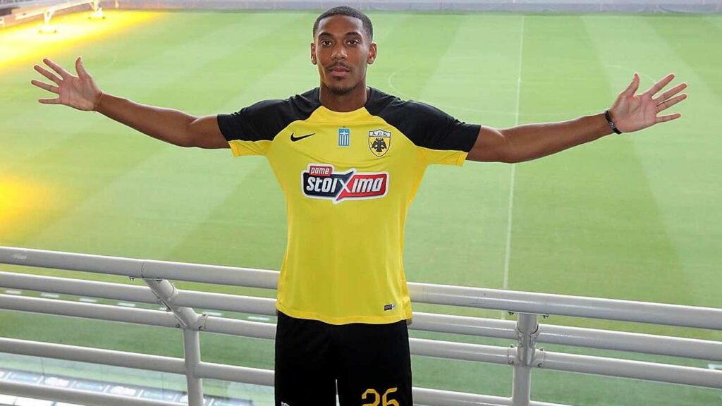 Anthony Martial in yellow and black soccer kit stands on a balcony with arms outstretched, a green pitch behind.