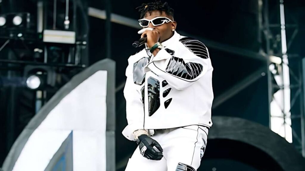 Antonio Brown in a futuristic white jacket with black paneling, white sunglasses, and gloves, mic in hand on a dark stage.