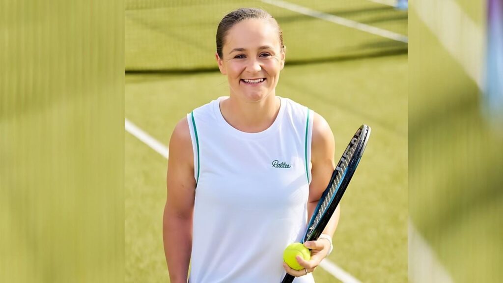 Ashleigh Barty in a white sleeveless tennis top with green trim stands on a sunny grass court, holding a ball and racket.