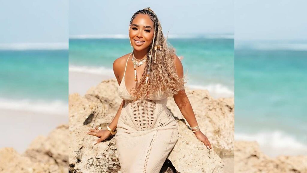 B Simone on sunlit beach wears a cream crochet dress, braided blonde hair and layered jewelry, turquoise water behind.