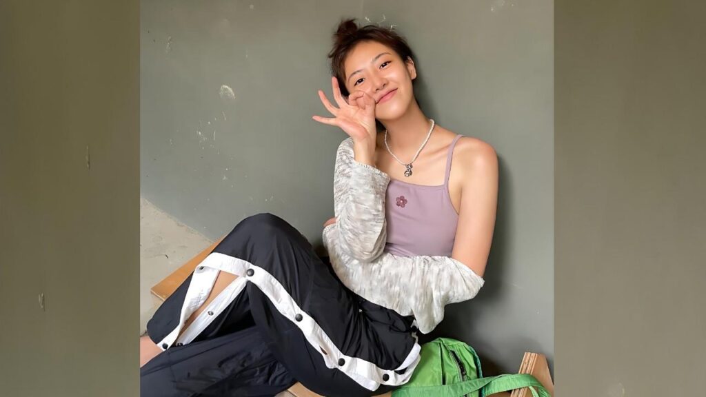 Bae Da-bin sits beside a green bag against a gray wall, smiling in a lilac tank top with a cardigan.