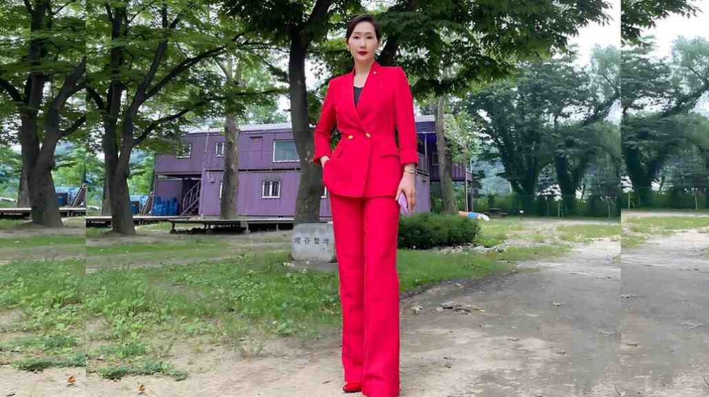Bae Hae-sun in a bright pink double-breasted suit with gold buttons stands in a park, holding a purple clutch.