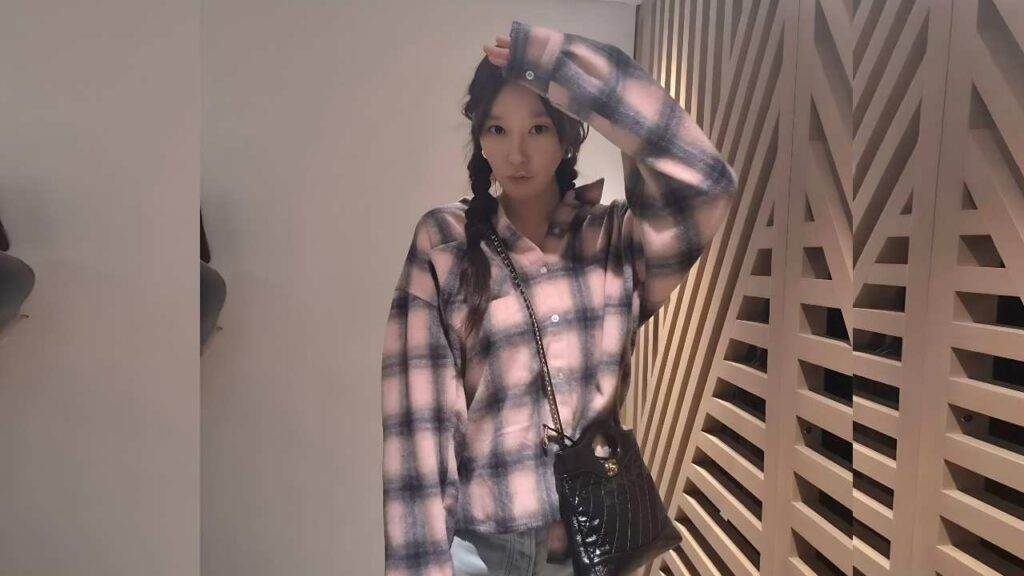 Bae Jin-ah in pink-gray plaid shirt with braided pigtails, raising her arm, standing beside a geometric wooden lattice wall with a crossbody bag.