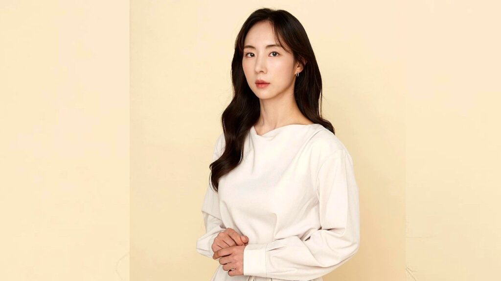 Bae So-eun with long dark hair wearing a white blouse stands against a pale beige backdrop, hands gently clasped at the waist.