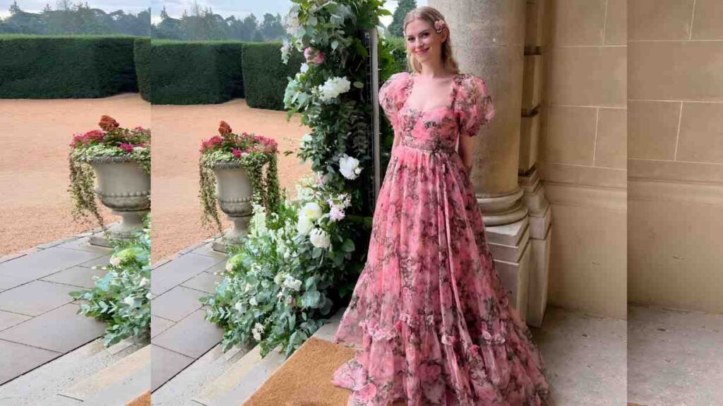 Barbara Dunkelman in a pink floral ballgown with a layered skirt poses beside a stone column, in a lush garden with planters behind her.