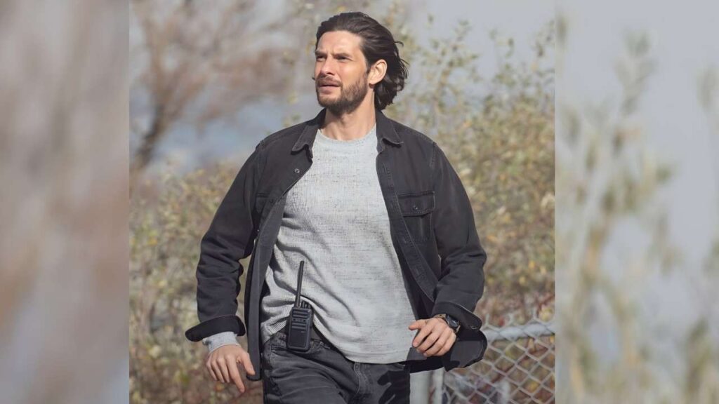 Ben Barnes with slicked-back hair and stubble walks in a black jacket over a gray sweater, radio clipped to his belt.