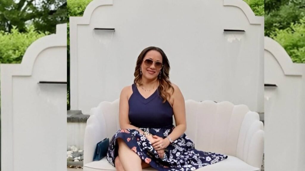 Beverly Heather in navy sleeveless top and floral skirt sits on a curved white outdoor bench, wearing sunglasses, in a garden setting.