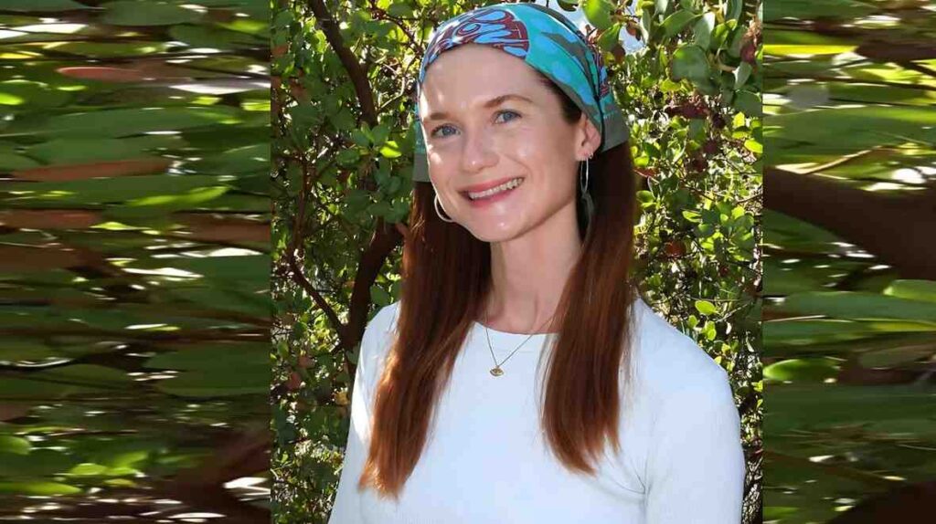 Bonnie Wright with long brown hair, colorful headscarf, hoop earrings, and white top, posed among sunny green foliage.