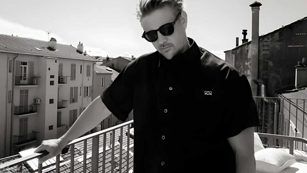 Boyd Holbrook in black short-sleeve shirt and sunglasses leans on a railing, rooftop and white buildings behind.