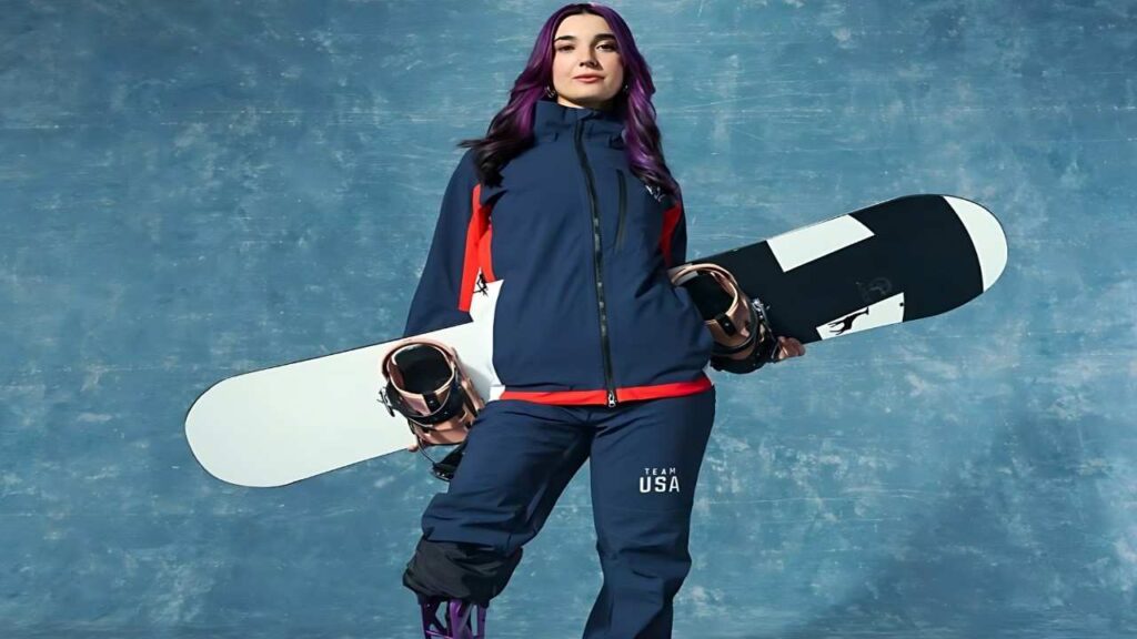 Brenna Huckaby in navy Team USA snowsuit stands against a blue backdrop, holding two snowboards across her torso.