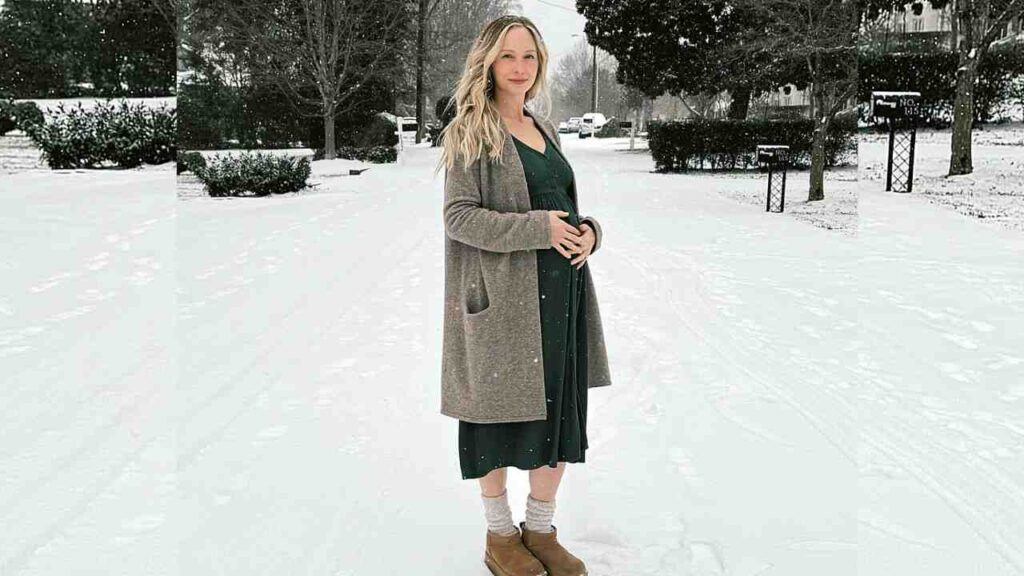Candice Accola stands in a snowy street, wearing a green dress with a long beige coat, cradling her belly as gentle snow falls.