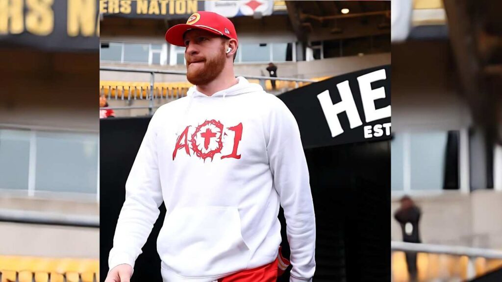 Carson Wentz in a white hoodie with a red A01 graphic, red cap, and white wireless earbuds walking through a stadium concourse.