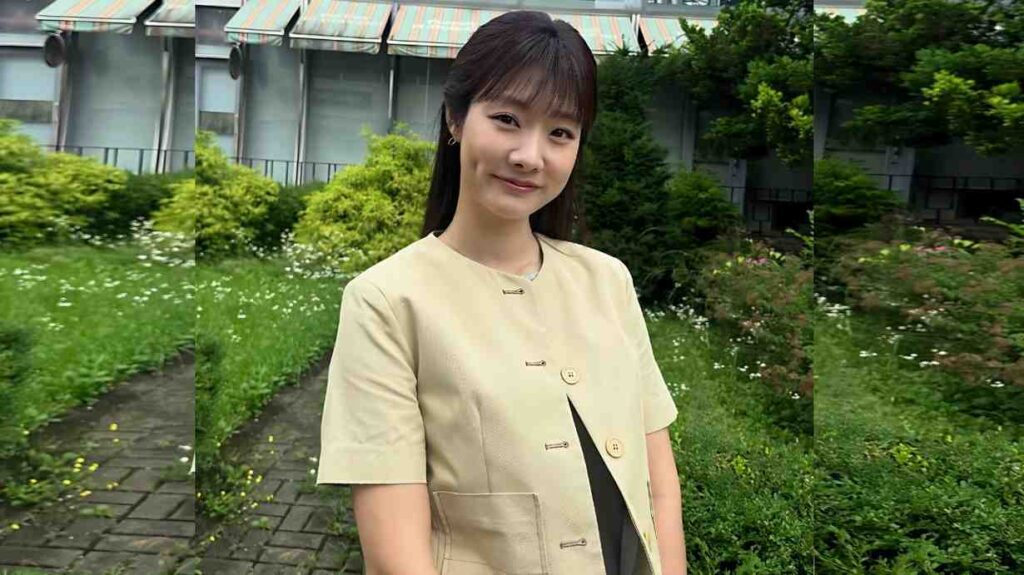 Cha Min-jee with dark hair in a beige short-sleeve jacket, standing in a lush garden with flowers and a building backdrop.