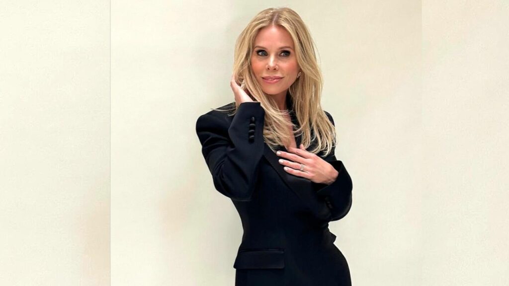 Cheryl Hines with long hair in a black tailored blazer stands against a pale wall, touching her hair and chest.i