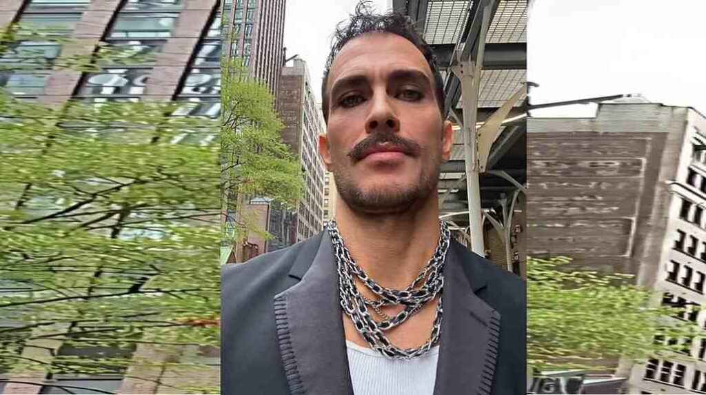 Cheyenne Jackson wth a trimmed mustache wearing layered silver chains over a dark blazer, posed outdoors in a city with trees.